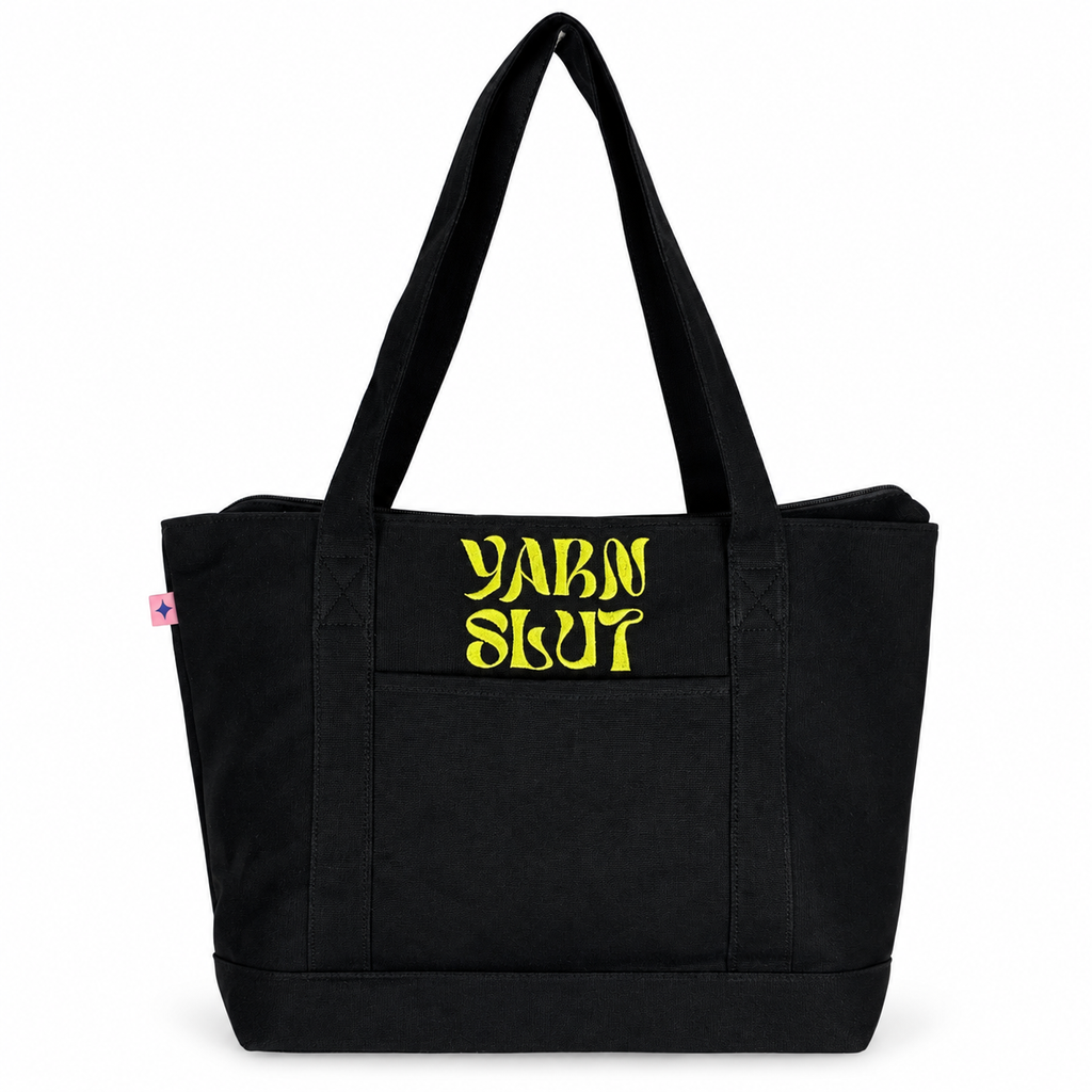 A black canvas tote bag with two shoulder straps and a front pocket, featuring the words 'YARN SLUT' printed in a stylized yellow font on the front pocket. A small pink tag is visible on the left side seam.