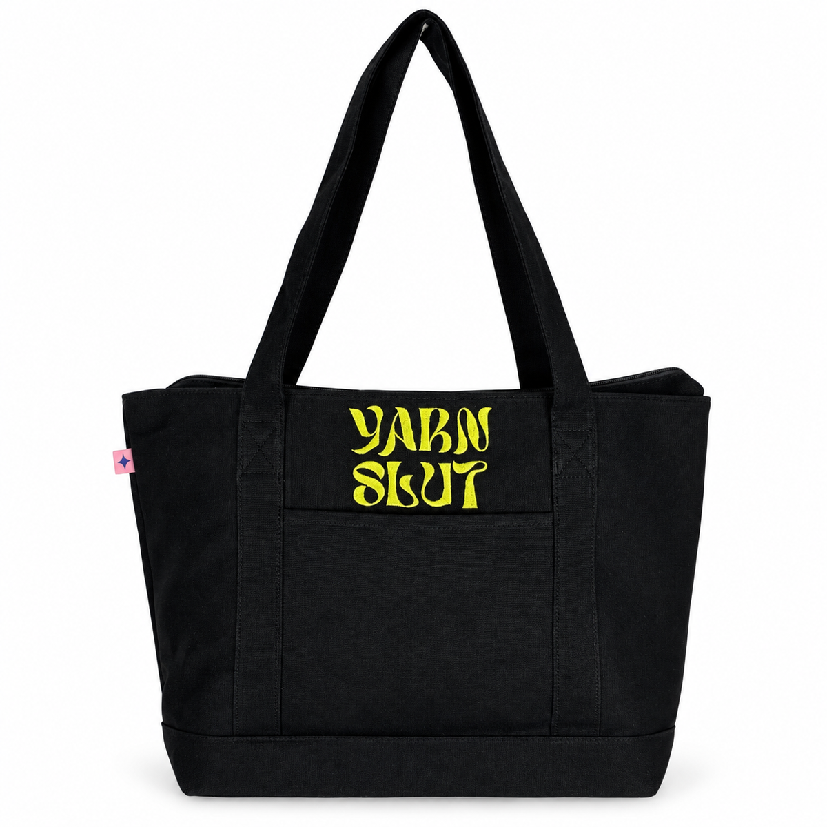 A black canvas tote bag with two shoulder straps and a front pocket, featuring the words 'YARN SLUT' printed in a stylized yellow font on the front pocket. A small pink tag is visible on the left side seam.